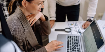 Businesswoman using laptop at office desk surrounded by colleagues, collaborating on project.
