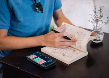 A man in a blue shirt uses a smartphone and book for studying indoors on a table.