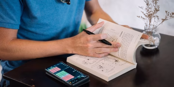 A man in a blue shirt uses a smartphone and book for studying indoors on a table.