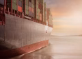 A colossal cargo ship loaded with containers navigates through calm waters against a vibrant sunset sky.