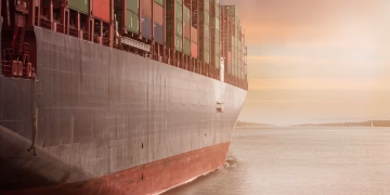 A colossal cargo ship loaded with containers navigates through calm waters against a vibrant sunset sky.