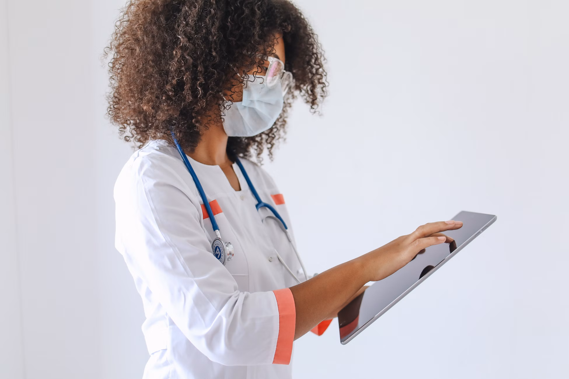 Female doctor with curly hair in lab coat using tablet, wearing a mask.
