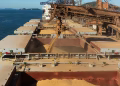 Aerial view of a cargo ship loading grain at a bustling industrial port by the sea.