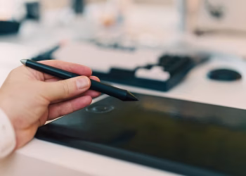 Close-up of a hand using a stylus on a digital tablet in an office setting.