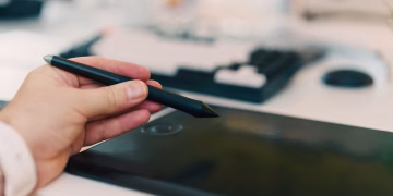 Close-up of a hand using a stylus on a digital tablet in an office setting.