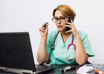 A woman in medical scrubs working with a laptop and phone, illustrating modern healthcare communication.