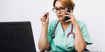 A woman in medical scrubs working with a laptop and phone, illustrating modern healthcare communication.