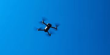 A black drone flying high in a clear blue sky, showcasing modern technology and flight capabilities.
