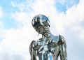A sleek chrome robot sculpture stands against a bright blue sky background.