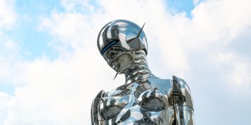A sleek chrome robot sculpture stands against a bright blue sky background.