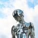 A sleek chrome robot sculpture stands against a bright blue sky background.