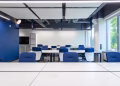 Various white desks with blue chairs placed in modern spacious workspace with big window and monitor in contemporary business center