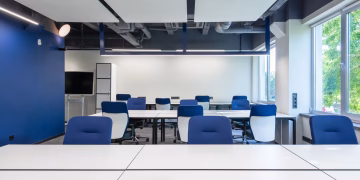 Various white desks with blue chairs placed in modern spacious workspace with big window and monitor in contemporary business center