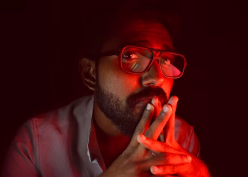 A thoughtful man with red ambient lighting, wearing glasses and a beard, looking upwards.