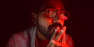 A thoughtful man with red ambient lighting, wearing glasses and a beard, looking upwards.