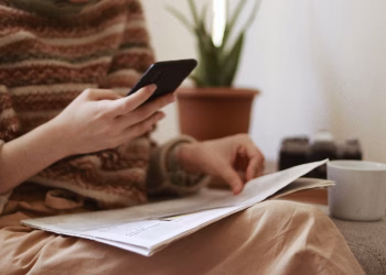 Crop unrecognizable female with newspaper text messaging on cellphone while sitting in armchair with cup of hot drink in house