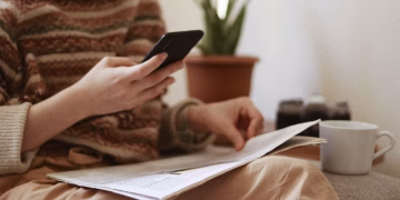 Crop unrecognizable female with newspaper text messaging on cellphone while sitting in armchair with cup of hot drink in house