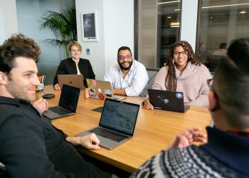 four people all on laptops, two men and two women, listen to person talking in a board meeting