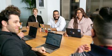 four people all on laptops, two men and two women, listen to person talking in a board meeting