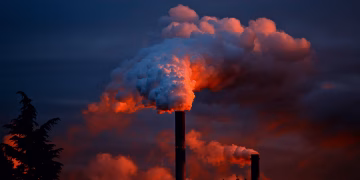 Smoke billows from industrial chimneys at sunset, highlighting pollution against a vibrant sky.