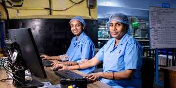 Two female workers at a textile factory operating computers in an industrial setting.
