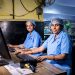 Two female workers at a textile factory operating computers in an industrial setting.