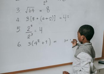 Side view of concentrated African American pupil standing near whiteboard in classroom and solving math examples during