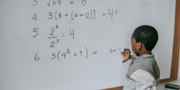 Side view of concentrated African American pupil standing near whiteboard in classroom and solving math examples during