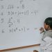 Side view of concentrated African American pupil standing near whiteboard in classroom and solving math examples during