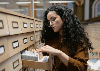 Curly haired woman in brown browsing a library card catalog with focus and curiosity.