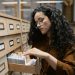 Curly haired woman in brown browsing a library card catalog with focus and curiosity.