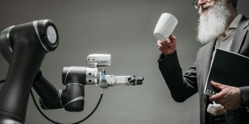 A bearded engineer holds a ceramic cup while a robotic arm assists, showcasing modern technology.