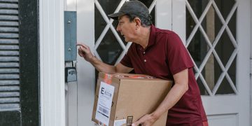 Man delivering a package at a front door, wearing a red shirt and cap.