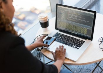 Black woman programming on a laptop with coffee, smartphone, and glasses on a desk in an office.