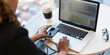 Black woman programming on a laptop with coffee, smartphone, and glasses on a desk in an office.
