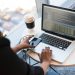Black woman programming on a laptop with coffee, smartphone, and glasses on a desk in an office.