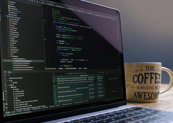 Close-up of laptop with coding software and a motivational coffee mug on a desk.
