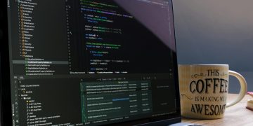 Close-up of laptop with coding software and a motivational coffee mug on a desk.