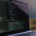 Close-up of laptop with coding software and a motivational coffee mug on a desk.