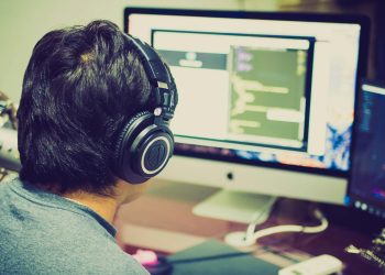 A programmer with headphones focuses on coding at a computer setup with dual monitors.
