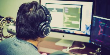 A programmer with headphones focuses on coding at a computer setup with dual monitors.