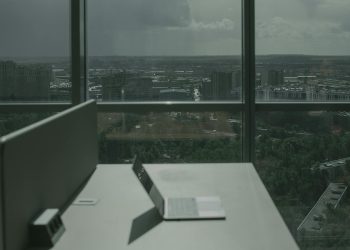 Laptop on office desk with a city skyline through a large window, offering a modern workspace view.