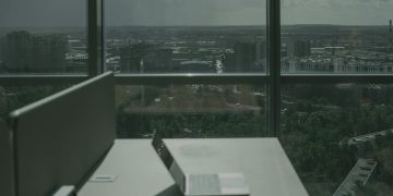 Laptop on office desk with a city skyline through a large window, offering a modern workspace view.