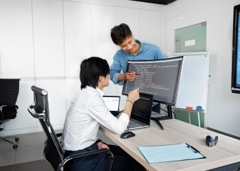 Two young professionals collaborating at a desk with laptops and monitors in a modern office setting.