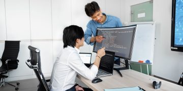 Two young professionals collaborating at a desk with laptops and monitors in a modern office setting.