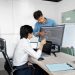 Two young professionals collaborating at a desk with laptops and monitors in a modern office setting.
