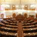 Wide view of an ornate legislative chamber with empty seats and chandeliers.