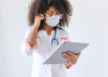 An African American female doctor wearing glasses and a face mask uses a tablet inside a medical facility.