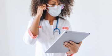 An African American female doctor wearing glasses and a face mask uses a tablet inside a medical facility.