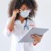 An African American female doctor wearing glasses and a face mask uses a tablet inside a medical facility.